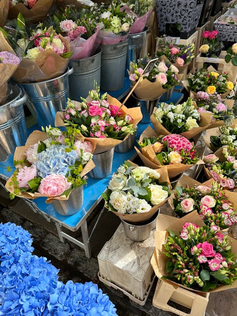 A top view of a florist arranging a bouquet of fresh flowers on a grey work table in a bright room. The bouquet includes pink roses, white ranunculus, and purple lilacs, with some stems and partially 