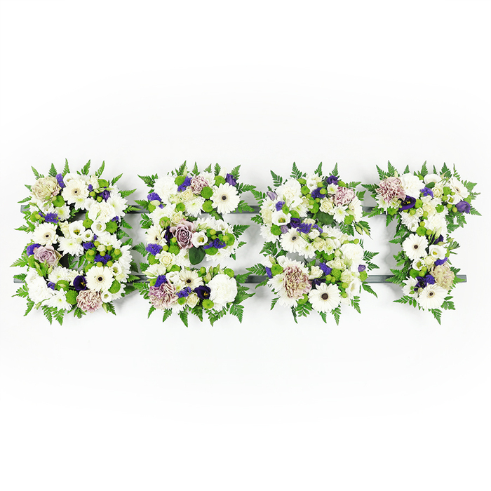 An overhead, horizontal composition spells out the word 'BEST' in full capital letters, each formed from a dense, carefully balanced collection of fresh blooms and verdant greenery. The design favours a serene palette of pristine whites and soft lavender-mauve accents: large, immaculate white Gerbera daisies with their dark centres create bold focal points, partnered with full white carnations and delicate lisianthus or chrysanthemum blooms that provide varied petal shapes and a soft, cloud-like texture. Interspersed are mauve and lavender roses and small deeper-purple filler clusters that add a quiet dignity and depth to the arrangement. Each letter is encircled by feathery fern fronds that give the edges a natural, sculpted softness while compact green button chrysanthemums punctuate the interiors with fresh, rounded bursts of green. The flowers are arranged upon a subtle dark grey or black base that peeks through at the edges, lending structure and clarity against the bright white photographic background. The overall effect is both articulate and tender-the floral typography reads clearly while remaining full of gentle movement and scent. As a florist based in Primrose Hill, Florist Primrose Hill often crafts these personalised sympathy letters for local memorials, wakes and funeral services around Chalk Farm and Regent's Park, mindful of the care such moments require. The composition feels like a quiet statement of honour and lasting remembrance, made to be seen up close and felt by those standing near the service.