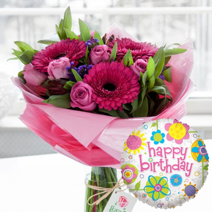 A radiant birthday bouquet and balloon pairing fills the frame with cheerful colour, evoking the feeling of a surprise delivery to a Primrose Hill flat on a bright morning. At the centre of the composition is a tall, cylindrical glass vase, its transparency showing the carefully trimmed flower stems gathered together in clear water. Rising from the vase is a generous arrangement dominated by large Gerbera daisies in shades of rich fuchsia and vivid magenta, their petals perfectly spaced around velvety dark centres that almost appear black, giving the bouquet a bold and contemporary edge. Surrounding these statement blooms are numerous roses in soft blush pink and deeper rose tones, some in tight buds that feel full of promise, others part-way open to reveal plush, layered petals that suggest classic romance. Between the larger flowers, tiny clusters of deep purple filler blooms, likely statice or a similar variety, create little pockets of shadow and texture, intensifying the depth of colour. A collar of fresh green foliage frames the entire arrangement, with smooth, bright leaves that fan out to create a rounded, abundant shape reminiscent of a walk through nearby Primrose Hill Park when everything is in bloom. Around the neck of the vase, pale pink, translucent cellophane is artfully gathered, forming a soft halo that echoes the pink tones of the flowers and rustles slightly in the light. Tied with rustic twine, a small off-white gift tag bearing a delicate rose motif hangs casually from the front, giving the sense of a thoughtfully chosen, premium birthday gift that might easily have been ordered from Florist Primrose Hill for same-day delivery across the neighbourhood. In the foreground, slightly to the right, stands a shiny, round foil balloon. Its white background is gently speckled with pale grey, overlaid with whimsical, colourful floral and circular patterns in bright yellow, lime green, sky blue, pink and deep purple. Across the centre flows the phrase "happy birthday" in playful pink cursive script, clearly announcing the occasion. The softly blurred backdrop, likely an airy living space or café near Regent's Park Road, keeps all attention on the joyful contrast of vivid blooms and festive balloon, capturing the warmth and celebration of a special birthday moment.