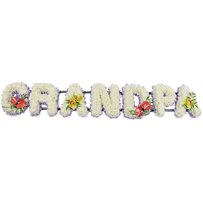 The photograph captures a personalised funeral flower tribute spelling out "GRANDPA" in solid, blocky capital letters that stretch in a single horizontal line. Each letter is meticulously covered with dense clusters of white chrysanthemums, the blooms positioned so closely together that they form a smooth, plush surface, like a soft quilt of petals symbolising purity, peace, and comfort for grieving hearts. Along the base of every letter, a delicate strip of lavender ribbon is just visible, adding a pale violet halo that outlines the forms and introduces a calm, soothing tone, similar to the soft evening light over Primrose Hill. Behind the lush floral faces, dark green framework connects the letters, allowing the tribute to be moved and displayed securely at a funeral, memorial service, or graveside gathering in the local area. Accents of vivid colour punctuate the white: on the 'G' and the 'D', rich clusters of deep red anthuriums sit proudly, their glossy, heart-shaped petals and pronounced spadices peeking out from a bed of dark green foliage and tiny purple fillers, expressing deep affection and heartfelt remembrance. The first and last 'A' are adorned with sprays of yellow-green orchids, their elegant, open blooms and gently curving stems trailing softly over the chrysanthemums, adding movement and a feeling of living grace. The tribute appears freshly made, as though arranged that day by a skilled Primrose Hill florist for same-day delivery to a chapel near Regent's Park Road or along the canal. This "GRANDPA" lettering is a thoughtful, bespoke memorial piece, offering families a beautiful, tactile way to honour a beloved grandfather's life and legacy.