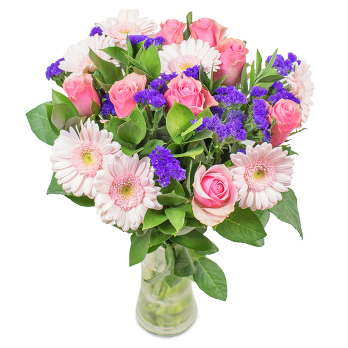 A precisely arranged floral bouquet capturing the spirit of Primrose Hill, showcased in a tall, clear glass vase against a pure white backdrop. The focal point comprises seven to nine pink roses, some recently arrived and elegantly curled, others just beginning to reveal their spiral formation. Five to seven larger gerbera daisies unfold pale pink petals from pale, creamy soft centres, echoing the subtle gradients of a Primrose Hill sunset. Scattered throughout are rich clusters of purple statice, offering bold contrast and tactile variance, reminiscent of the playful vibrancy seen at local boutique markets. Emerald foliage envelopes the lower stems, layering depth and freshness that feels synonymous with the greenery lining Gloucester Avenue. Visible stems and water inside the vase reinforce a sense of natural, hand-tied abundance. Suitable as a memorable gift for special occasions or simply to brighten a home or workspace near Chalk Farm, this bouquet radiates festive joy and diligent artistry-an emblem of care and celebration unique to Primrose Hill.